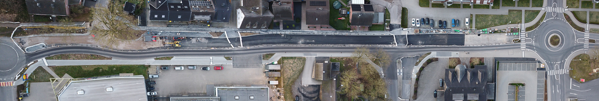 Eine lange Straße ist zu sehen, die jeweils an beiden Enden mit einem Kreisverkehr abschließt. Umbaumaßnahmen werden auf dem Bild deutlich. Zahlreiche Fahrzeuge und Gebäude sind an der Straße platziert. Außerdem befinden sich einige Grünstreifen und Bäume im näheren Umfeld zur Straße.